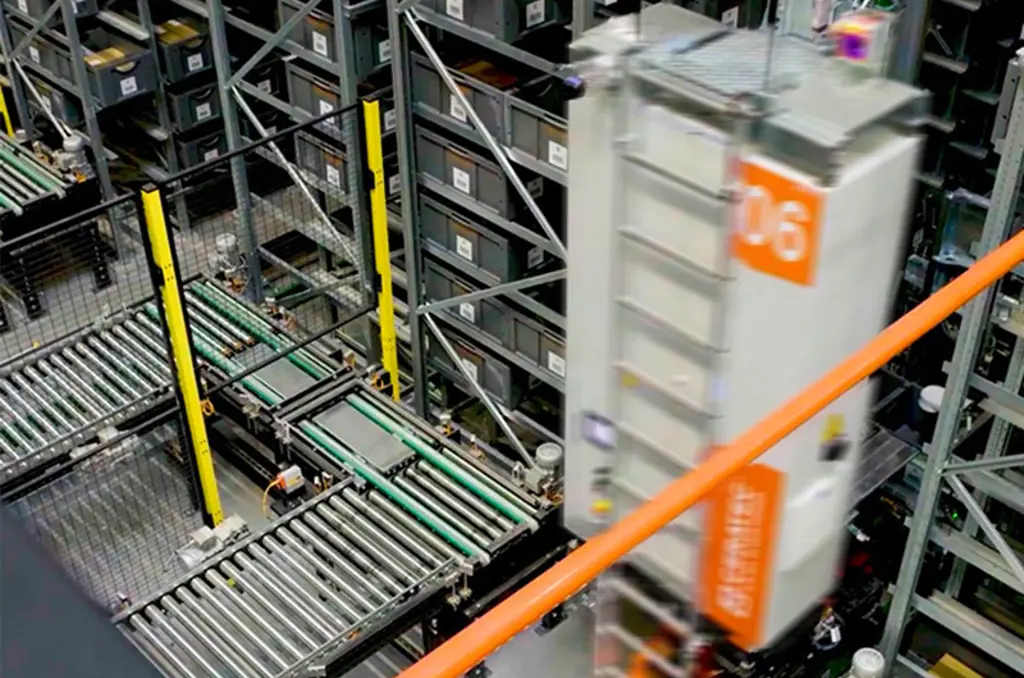 Magazzino automatizzato con nastri trasportatori e robot di stoccaggio ad alta velocità che si muove tra alte scaffalature di contenitori.